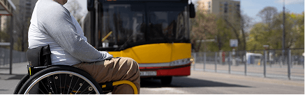 Person using a wheelchair waiting at a bus stop with a bus approaching in the background.