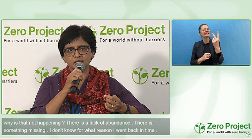 a woman speaks at a Zero Project event, accompanied by a sign language interpreter and live subtitles.
